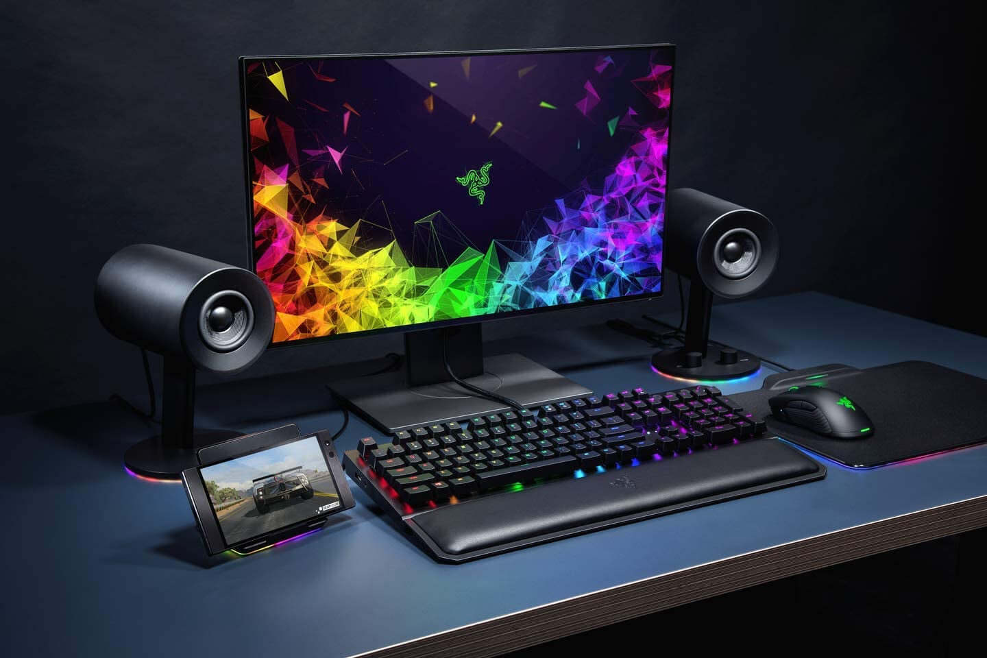 Gaming setup featuring Razer Nommo Chroma speakers, keyboard, mouse, and vibrant monitor display.